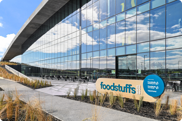 Foodstuffs' entrance. It is a large building with modern architecure with tall reflective windows. A minimalistic sign with the company logo in the colors beige and light blue greet the visitor with the company name 'foodstuffs' in lowercase.